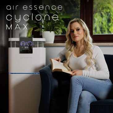 Kobieta o długich blond włosach siedzi na granatowym krześle z otwartą książką. Obok niej znajduje się Oczyszczacz powietrza z nawilżaczem Air Essence™ Cyclone Max. W tle zielona roślina i okno.
