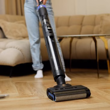 Osoba w dżinsach i białych skarpetkach używa odkurzacza Stojący Odkurzacz Myjący, Bezprzewodowy 7w1, Air Essence™ Typhoon Drive Jet na drewnianej podłodze w salonie z beżową sofą i szklanym stolikiem kawowym w tle.