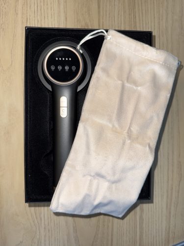 Click to view full screen Ręczne urządzenie elektroniczne z przyciskami i okrągłym wyświetlaczem znajduje się w czarnym pudełku obok kremowego, aksamitnego woreczka ze sznurkiem na powierzchni z jasnego drewna.